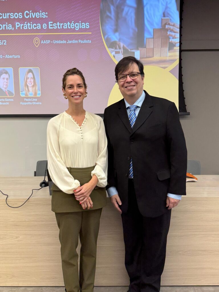 Foto com a presidente Paula Lima Hyppolito Oliveira e o Coordenador do Curso Gilberto Gomes Bruschi na Unidade Jardim Paulista. Em frente de uma mesa e um telão de apresentações